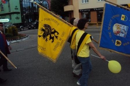 Uczestnik zjazdu Kaszubów niesie żółtą flagę z gryfem i balon na ulicy.