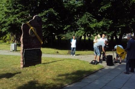 Drewniany diabeł kaszubski i ludzie na zjeździe w Pucku, otoczeni zielenią.