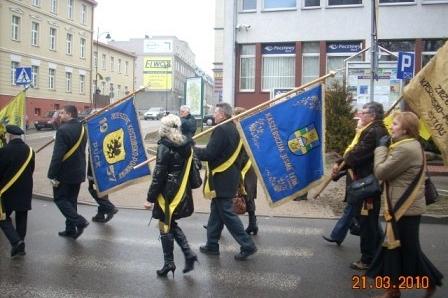 Procesja osób w mundurach z kaszubskimi flagami na ulicy w Kartuzach.