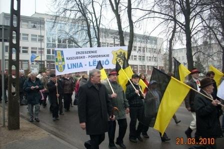 Uczestnicy Dnia Jedności Kaszubów maszerują z flagami i transparentem ulicami Kartuz.