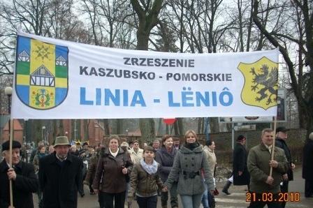 Procesja ludzi z banerem Linia-Leniō na tle drzew, symbolizująca jedność Kaszubów.