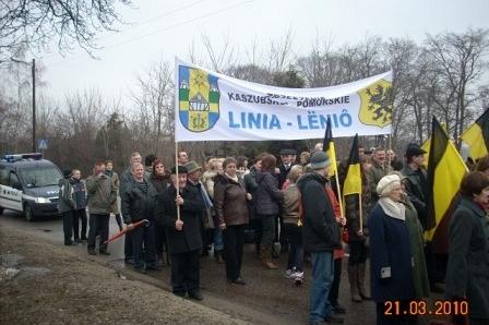 Pochód Kaszubów z żółto-czarnymi flagami i transparentem Lina. Lënô na Dzień Jedności.