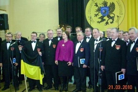 Eleganccy uczestnicy Dnia Jedności Kaszubów z flagą kaszubską na tle regionalnego emblematu.