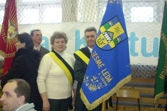 Mężczyzna trzyma kaszubską flagę, obok stoi kobieta, w tle okna i inni ludzie.