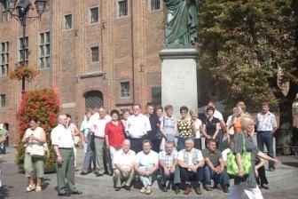 Grupa turystów pozuje na placu z zabytkowym budynkiem i pomnikiem w tle.
