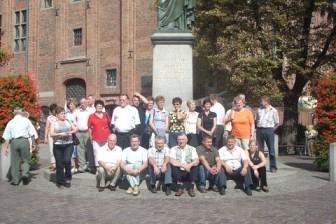 Uczestnicy wycieczki pozują przed pomnikiem na tle ceglanego budynku z czerwoną dachówką.
