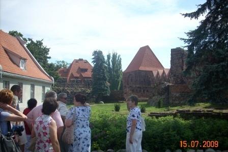 Uczestnicy wycieczki oglądają historyczne budynki z czerwonymi dachami otoczone zielenią.