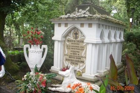 Biały, zdobiony grobowiec z kwiatami i urną w otoczeniu zieleni.