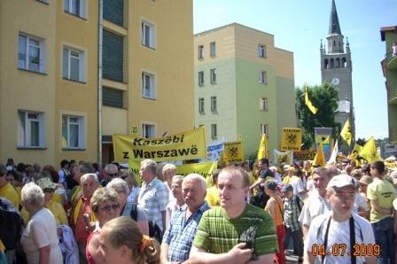 Tłum uczestników Zjazdu Kaszubów w Bytowie, z żółtymi flagami, budynkami i wieżą kościoła.