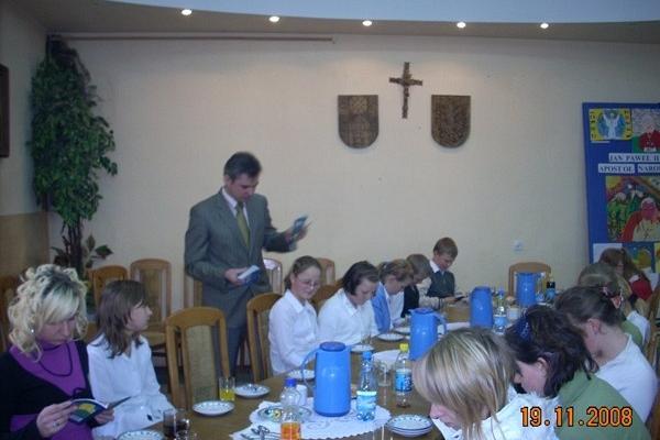 Mężczyzna prowadzi konkurs wiedzy dla młodzieży w sali z herbami.