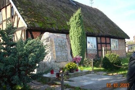 Szachulcowy dom ze strzechą, przed nim kamienny głaz wśród zieleni.