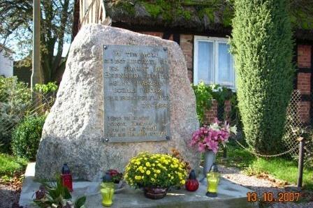 Pamiątkowy kamień w Szymbarku z bukietami kwiatów, oświetlony słońcem.