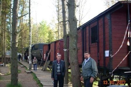 Dwóch mężczyzn na tle czerwonych wagonów kolejowych i drzew podczas wycieczki w Szymbarku.
