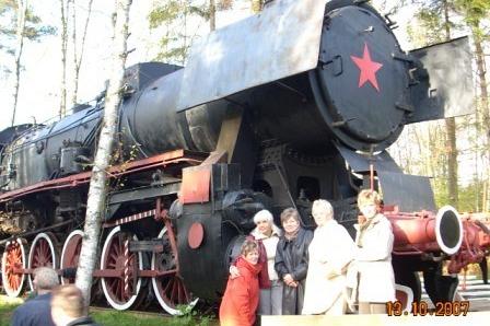 Uczestnicy wycieczki oglądają zabytkowy parowóz z czerwoną gwiazdą na tle zieleni.