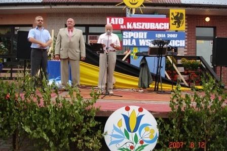 Trzech mężczyzn na festynie sportowym na Kaszubach, przy stoisku z banerem i herbem.