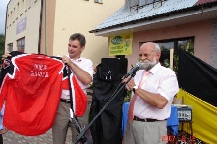 Dwaj mężczyźni prezentują czerwoną koszulkę sportową na festynie, obok mikrofonu.