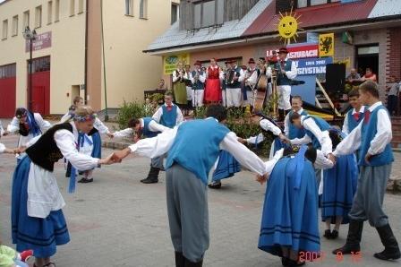 Grupa ludzi w tradycyjnych kaszubskich strojach tańczy w kręgu na zewnątrz, prawdopodobnie podczas festynu.