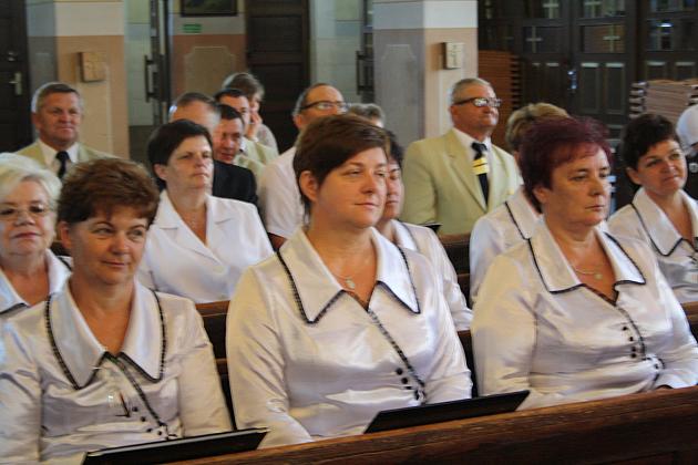 Grupa kobiet w jednakowych białych koszulach z ciemnymi kołnierzami siedzi w rzędach, patrząc przed siebie.