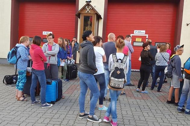 Grupa młodzieży z walizkami przed czerwonym budynkiem, początek kolonii letniej.
