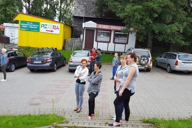 Uczestniczki kolonii idą przed budynkiem z żółtym pojazdem i autami w tle.