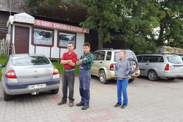 Trzech mężczyzn stoi przed budynkiem i samochodami w Karpaczu, prawdopodobnie na terenie kolonii.