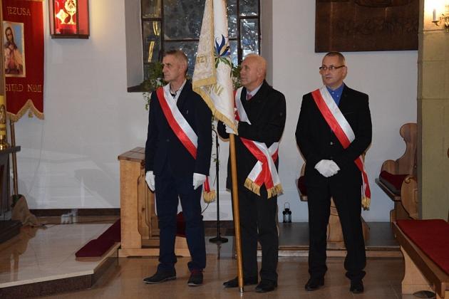 Trzech pszczelarzy w uroczystych strojach z flagą koła podczas spotkania.