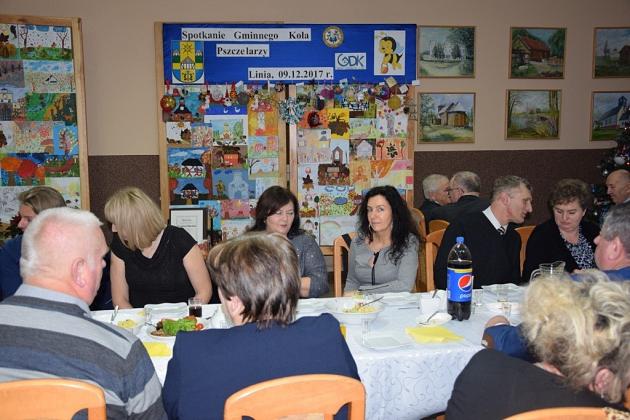 Uczestnicy świątecznego spotkania Gminnego Koła Pszczelarzy siedzą przy stołach.