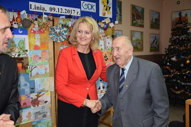 Kobieta w czerwieni wita starszego pszczelarza podczas świątecznego spotkania.