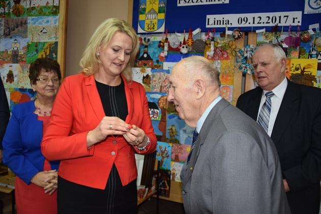 Młoda kobieta wręcza coś starszemu pszczelarzowi na świątecznym spotkaniu.