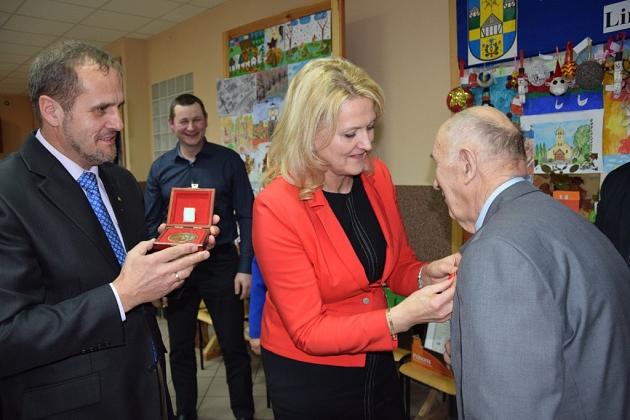 Kobieta wręcza odznaczenie starszemu panu, podczas gdy obok mężczyzna trzyma czerwoną nagrodę.
