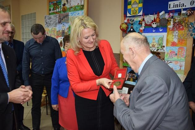 Kobieta w czerwonym żakiecie wręcza drobny upominek mężczyźnie na spotkaniu pszczelarzy.