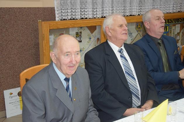 Trzech starszych pszczelarzy siedzi przy stole podczas świątecznego spotkania, jeden uśmiecha się.