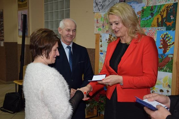 Kobieta w czerwonej marynarce wręcza upominek drugiej kobiecie, mężczyzna stoi w tle.