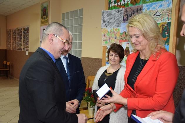Kobieta w czerwonej marynarce wręcza upominek mężczyźnie na spotkaniu pszczelarzy.