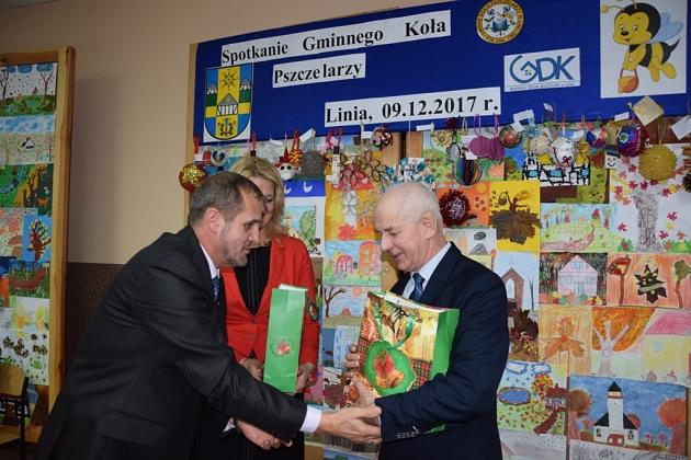 Dwóch mężczyzn wymienia uścisk dłoni i upominek na świątecznym spotkaniu pszczelarzy.