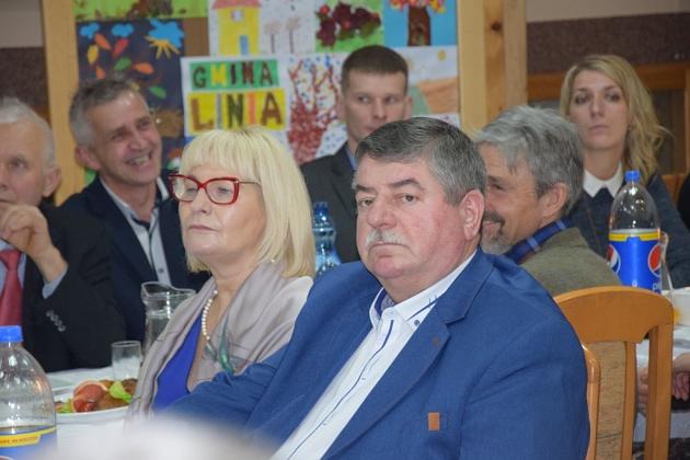 Mężczyzna w marynarce i inni uczestnicy świątecznego spotkania pszczelarzy przy stole.