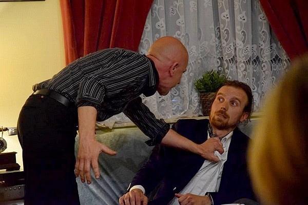Scena teatralna: aktor w ciemnej koszuli chwyta drugiego za ubranie w dramatycznej pozie.